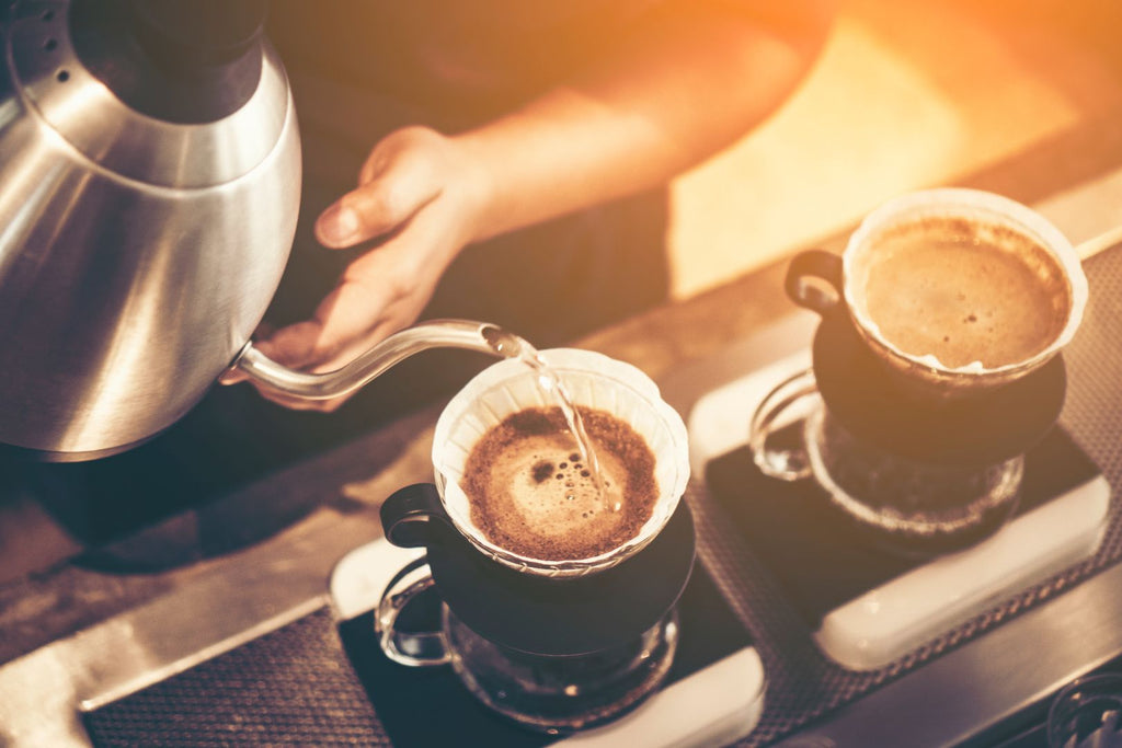 A close-up of a person pouring filtered water from a kettle into a coffee strainer. Two coffee cups sit on the counter.